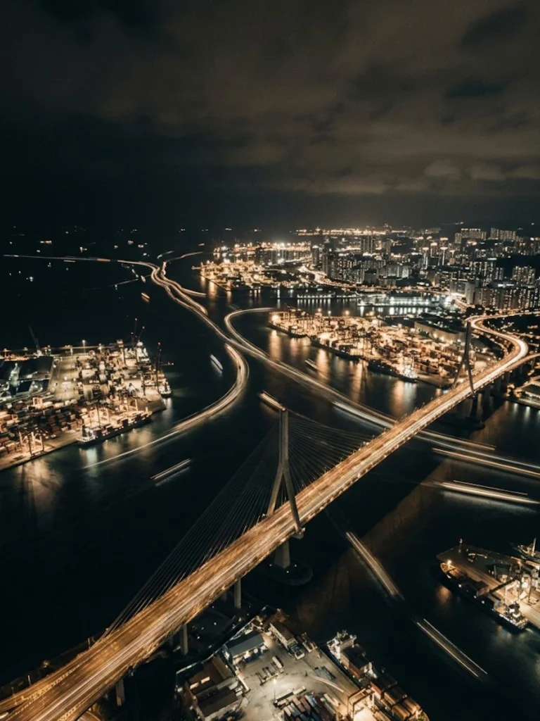 Night aerial view of a major port: illuminated cable-stayed bridge, container terminals, and city lights reflecting on dark water